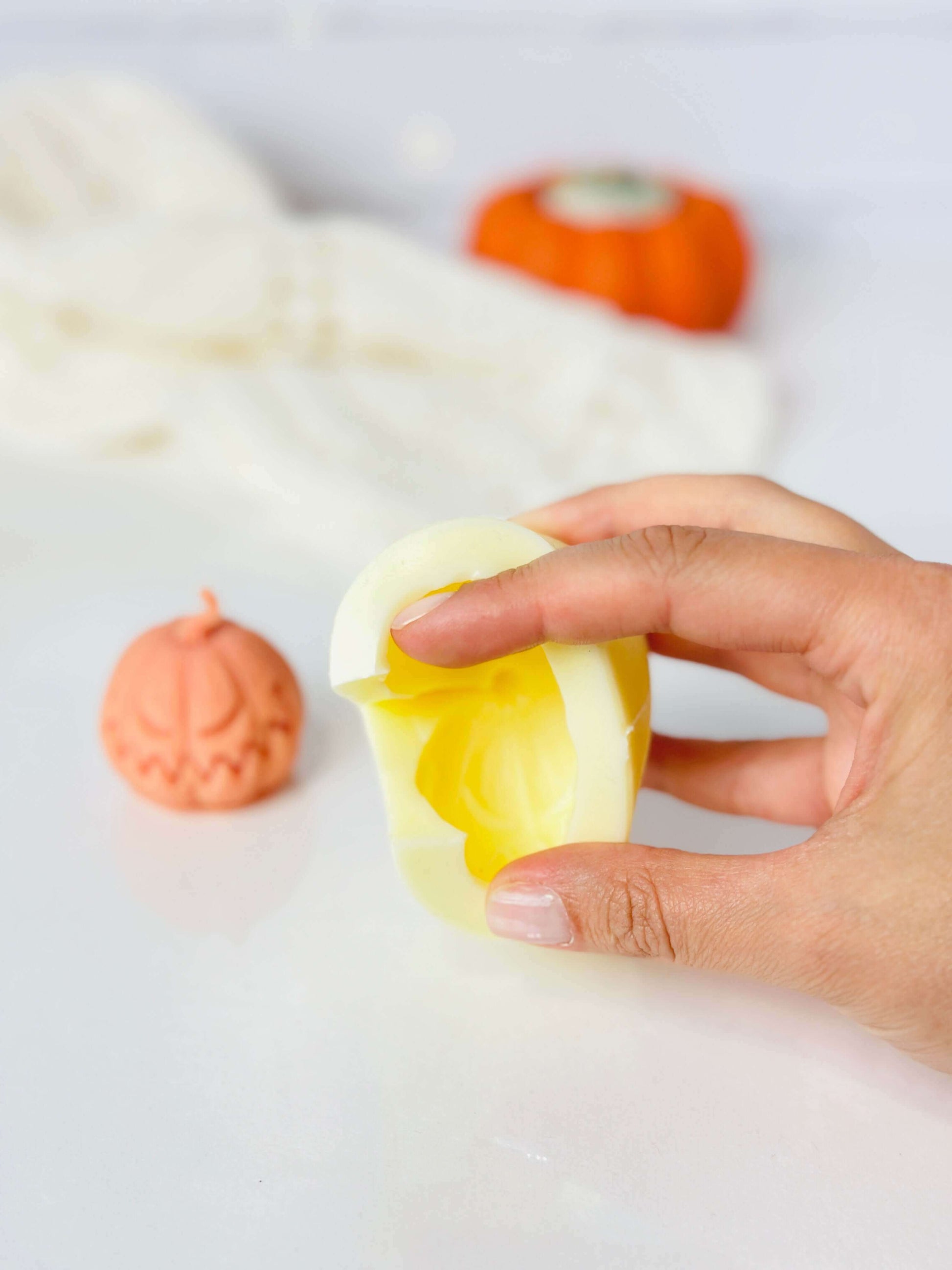 Hand holding a yellow candle mold while a pumpkin-shaped candle sits nearby, showcasing Halloween creativity.