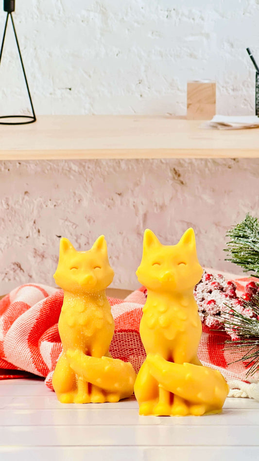 Two vibrant yellow fox-shaped candles on a wooden table with a cozy background, showcasing handmade elegance.