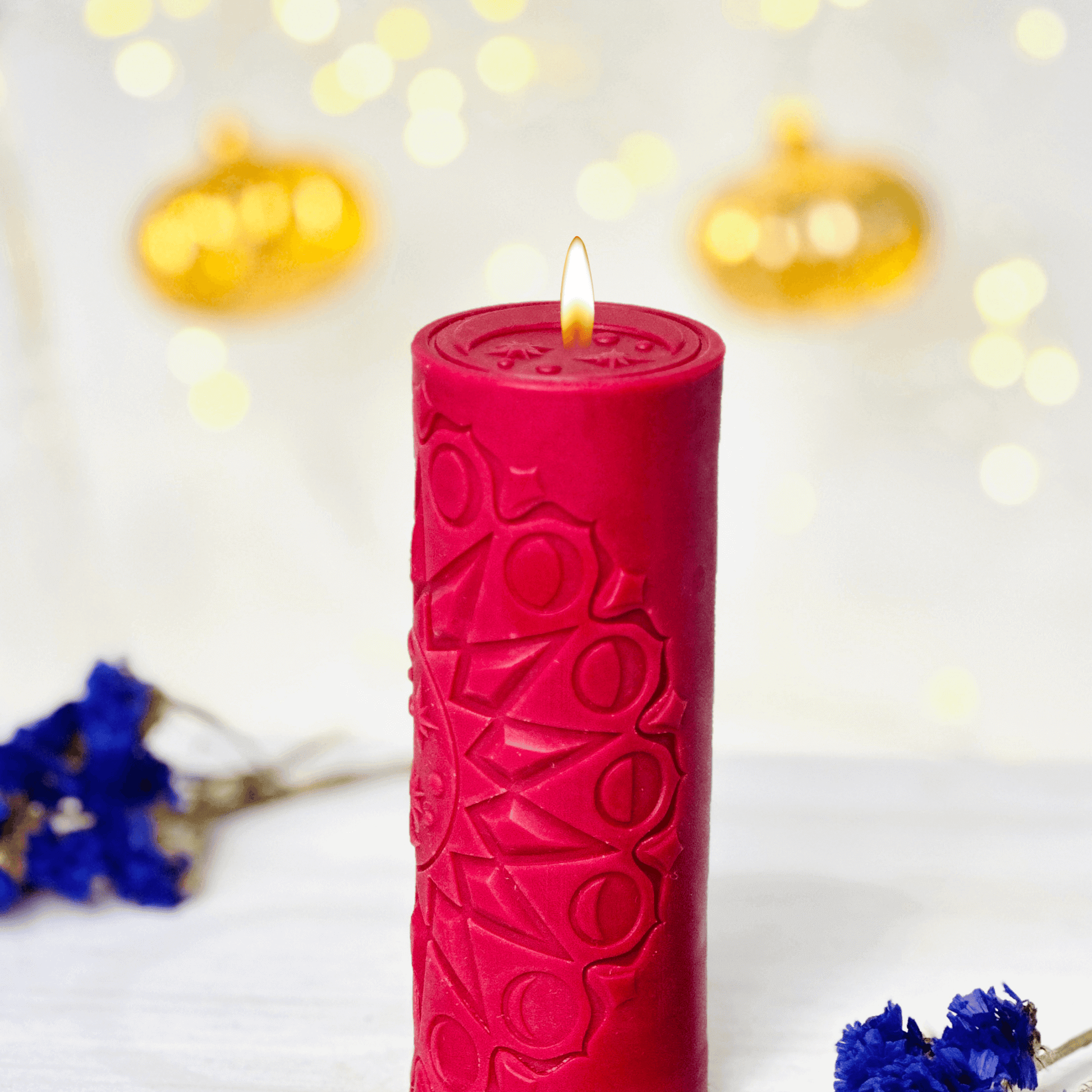 Red spiritual candle with intricate design, glowing wick, surrounded by blue flowers and festive lights.